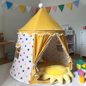May include: A yellow and white play tent with a star pattern and pom-pom trim. The tent has a circular base and a conical roof. A large yellow sun-shaped pillow is inside the tent. Colorful triangle flags hang in the background.