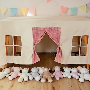 May include: A cream-colored play tent with a red and white checkered curtain and window cutouts. A row of plush teddy bears in various colors and styles are lined up in front of the tent. A colorful banner hangs above the tent.