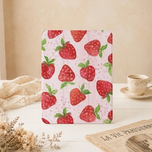 Könnte beinhalten: Eine rechteckige Tablethülle mit einem sich wiederholenden Muster aus roten Erdbeeren mit grünen Blättern auf einem hellrosa Hintergrund mit kleinen rosa Sprenkeln. Die Hülle hat eine glatte Oberfläche und eine sichtbare vertikale Falzlinie.