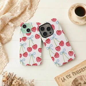 May include: Two white phone cases with a repeating pattern of red cherries, green stems, and light blue bows. The cases have camera cutouts and are placed on a white surface. One case has a mint green camera module.