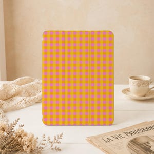 Könnte beinhalten: Eine rechteckige Tablethülle mit einem lebendigen Vichy-Karomuster aus abwechselnden Quadraten in leuchtendem Pink und Goldgelb. Die Hülle hat eine glatte, flache Oberfläche mit vertikalen Faltlinien, die auf der Vorderseite sichtbar sind.