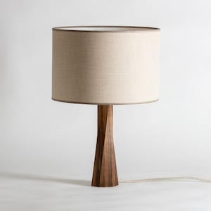 May include: A wooden table lamp with a beige fabric shade. The lamp has a dark brown, geometric base and a neutral-colored lampshade. The lamp is plugged into a white wall. The lamp is a decorative lighting fixture.