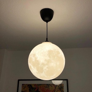 Peut inclure: Une lampe suspendue sphérique ressemblant à la lune, avec une surface texturée et une lumière chaude et brillante. La lampe est suspendue à un luminaire noir fixé au plafond. Le diamètre de la lampe est d'environ 30 cm.