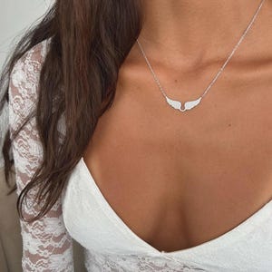 Peut inclure: Un collier argenté avec un pendentif en forme d'ailes. Le pendentif est orné de petites pierres scintillantes. Le collier est porté sur le cou d'une personne, sur un haut en dentelle blanche.