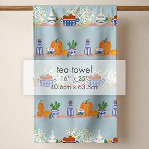 May include: A light blue tea towel measuring 16 inches by 25 inches, featuring a repeating pattern of cats, orange pumpkins, blue and white striped bottles, potted green plants, and small rectangular tins labeled Azul, arranged on horizontal shelves.