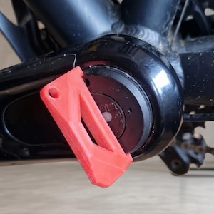 Può includere: Primo piano dell'area del movimento centrale di una bicicletta. Una copertura protettiva rossa brillante, stampata in 3D, è attaccata alla guarnitura nera. Il telaio della bici è nero e lo sfondo è una superficie marrone chiaro.