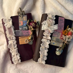 Könnte beinhalten: Zwei dunkelbraune Leder-Notizbücher, verziert mit weißer Spitzenborte, getrockneten Blumensträußen und Vintage-Konzerttickets. Jedes Notizbuch ist mit einer Metallklammer, Anhängern und kleinen Papier-Ephemera auf den Vorderseiten versehen.