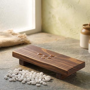 May include: A rectangular, wooden soap dish with a dark brown finish, supported by two small legs. Several water droplets are visible on the surface. A pile of small, white stones is in front of the dish. The background includes a window and a ceramic jar.