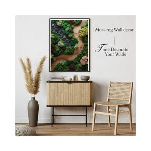 May include: A framed moss rug wall decor piece with a black frame, featuring a natural landscape design with various shades of green, brown, and beige. The image is displayed above a light wood cabinet with black legs, a gray vase, and a woven chair.