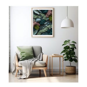 Op de afbeelding: Een ingelijste print met een close-up van een natuurlijke scène met groene, roze en grijze tinten. Het kunstwerk hangt boven een grijze fauteuil met een groen kussen en een grijze deken. Een bijzettafel en een plant in pot zijn ook zichtbaar.