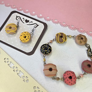 Puede incluir: Juego de joyas con temática de donuts, que incluye un par de pendientes y una pulsera. Los pendientes y la pulsera están adornados con dijes de donuts en miniatura en varios colores, como amarillo, marrón, negro y rosa. Las joyas se presentan sobre un fondo decorativo.