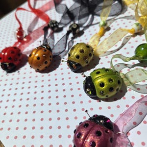 May include: A collection of colorful ladybug ornaments with decorative ribbons. The ladybugs are in various colors, including red, orange, gold, green, and pink, each with black spots and antennae. The ribbons are red, black, yellow, green, and pink.