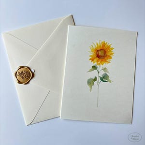 May include: A cream-colored greeting card featuring a watercolor illustration of a single yellow sunflower with green leaves, paired with a matching envelope sealed with a gold wax seal embossed with a rose design and the text Chapter & Bloom.