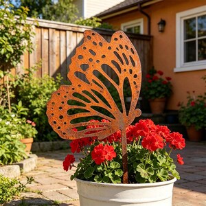 May include: A rust-coloured metal butterfly garden stake with intricate cut-out wing patterns. The butterfly ornament is positioned upright in a white planter filled with vibrant red geranium flowers, creating a decorative garden accent.