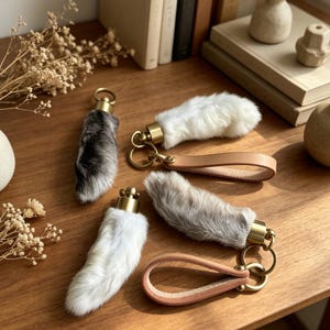 May include: Four faux fur keychain charms in white, gray, and black, with gold-tone hardware and leather straps. The charms are arranged on a wooden surface, with dried flowers and books in the background.