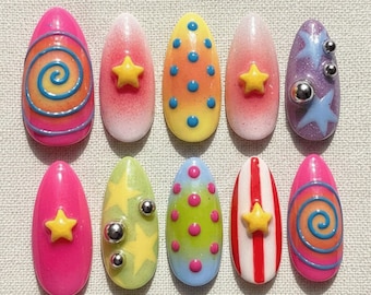 Peut inclure: Un ensemble de dix faux ongles aux motifs variés. Les ongles présentent des couleurs vives comme le rose, le jaune et le violet, avec des décorations telles que des étoiles, des spirales, des pois et des accents métalliques. Les ongles sont de forme ovale.
