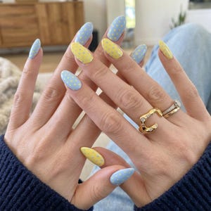 Puede incluir: Uñas en forma de almendra con base azul claro y lunares amarillos o base amarilla con lunares azul claro. En el dedo anular se llevan dos anillos dorados, uno con forma orgánica irregular y otro con una fina banda de piedras.