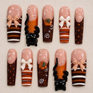 Op de afbeelding: Een set van tien kunstnagels met een nude basis en bruine en oranje accenten. De ontwerpen bevatten horizontale strepen, stippen, witte en perzikkleurige strikjes, 3D zwarte katten en kleine oranje pompoenen met witte kruldetails en pareltjes.