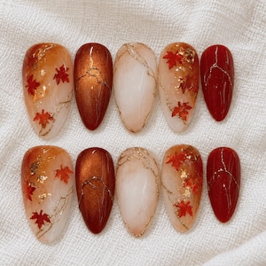 Op de afbeelding: Een set van tien amandelvormige press-on nagels met een herfstachtig ontwerp. De nagels hebben een mix van glanzend dieprood, metallic koper en doorschijnende crèmekleurige afwerkingen, geaccentueerd met gouden bladvlokken en gouden takjes.