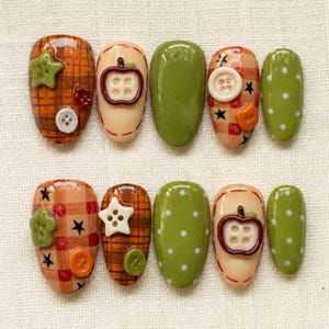 Op de afbeelding: Een set van tien kunstnagels met diverse ontwerpen, waaronder oranje en bruine ruitpatronen, effen olijfgroen en crèmekleurige achtergronden. De nagels zijn versierd met kleine plastic knoopjes in de vorm van sterren, cirkels en appels.