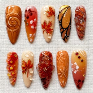 Op de afbeelding: Een set van tien amandelvormige press-on nagels met herfstgeïnspireerde ontwerpen in tinten oranje, rood, bruin en crème. De nagels tonen complexe 3D-texturen, bloemmotieven, esdoornbladeren, vlindervleugelmotieven en kleine strass-steentjes.
