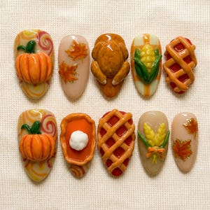 Op de afbeelding: Een set van tien kunstnagels met 3D-ontwerpen in het thema Thanksgiving. De collectie bevat gebeeldhouwde pompoenen, een gebraden kalkoen, maïskolven, taarten met een rasterpatroon, een toefje slagroom, tarwearen en oranje herfstbladeren.