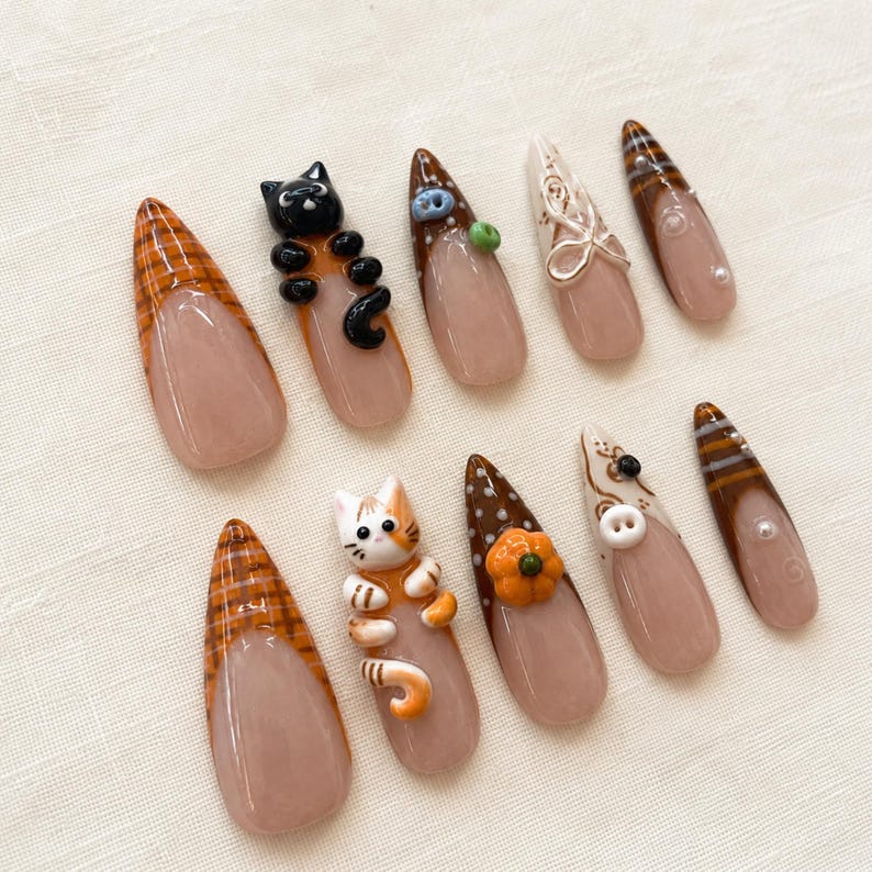 Op de afbeelding: Een set van tien kunstnagels met een nude basis en diverse herfstthema ontwerpen, waaronder 3D zwart-witte katfiguren, een kleine oranje pompoen, witte knopen, bruine ruitpatronen en bruine strepen met kleine witte parelaccenten.