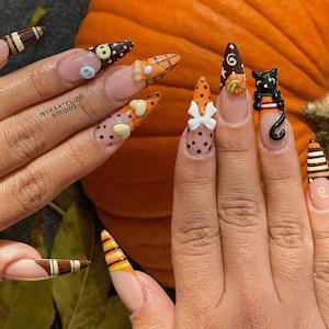 May include: Stiletto-shaped artificial nails featuring a clear base with various autumn-themed 3D designs including brown and orange polka dots, stripes, a black cat, a white bow, and pumpkin accents. The text MYRAATELIER STUDIO is printed on one nail.
