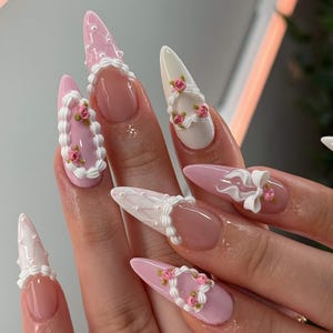 May include: Stiletto-shaped nails featuring a mix of soft pink and white polish. The designs include raised white piping, small pink rose accents, and tiny white pearls. Some nails have a French-style tip, while others are fully painted with intricate 3D detailing.