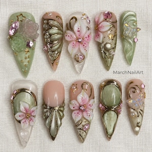 Op de afbeelding: Tien kunstnagels met complexe 3D-ontwerpen in saliegroen, zachtroze en goud. De versieringen bestaan uit gebeeldhouwde bloemen, metalen krullen, iriserende vlinders en kleine kristallen. De tekst MarchNailArt is zichtbaar op de afbeelding.