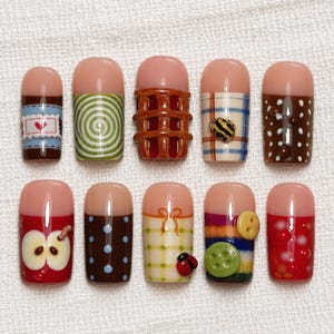 May include: A set of ten press-on nails featuring diverse designs including a red heart, green spiral, brown waffle grid, plaid with a bee, brown with white dots, apple slice, blue with dots, yellow bow, ladybug, buttons, and red berries.