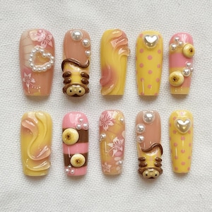 Könnte beinhalten: Ein Set aus zehn künstlichen Nägeln mit verschiedenen Designs. Die Nägel weisen eine Reihe von Farben auf, darunter Gelb, Rosa und Braun, mit dekorativen Elementen wie Perlen, floralen Mustern und Herzformen. Einige Nägel haben 3D-Verzierungen.