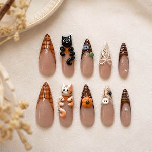 Op de afbeelding: Een set van tien amandelvormige plaknagels met een nude basis en diverse herfstthema-ontwerpen, waaronder een zwarte kat, een lapjeskat, een pompoen, een witte knoop, ruitpatronen, bruine stippen en kleine zilveren kraaltjes.