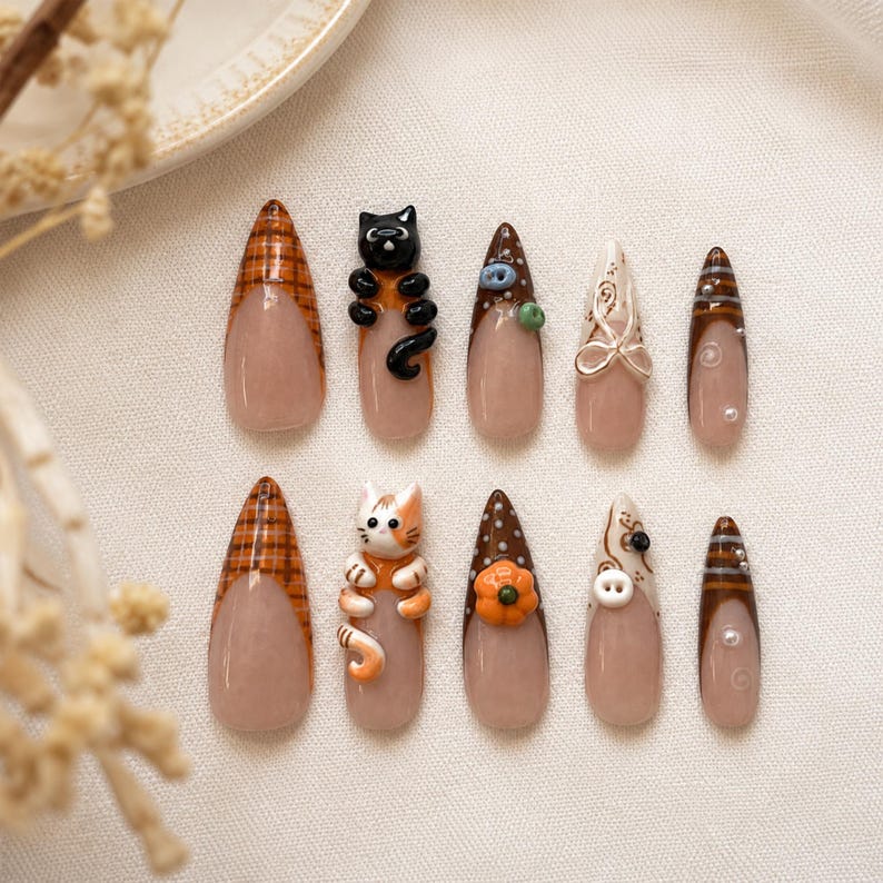 Op de afbeelding: Een set van tien amandelvormige plaknagels met een nude basis en diverse herfstthema-ontwerpen, waaronder een zwarte kat, een lapjeskat, een pompoen, een witte knoop, ruitpatronen, bruine stippen en kleine zilveren kraaltjes.