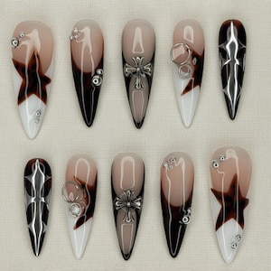 May include: A set of ten long, pointed artificial nails. Each nail features a unique design with a combination of colors including white, brown, and black. Some nails have silver embellishments, crosses, or starburst patterns. The nails are arranged on a neutral surface.
