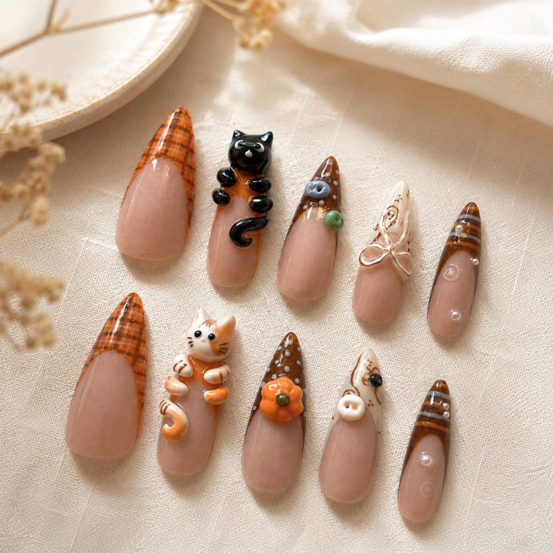 Op de afbeelding: Een set van tien amandelvormige kunstnagels met een nude basis en diverse herfstthema ontwerpen, waaronder oranje ruitpatronen, 3D zwarte en oranje katfiguren, een kleine oranje pompoen, witte stippen, bruine strepen en kleine witte parels.