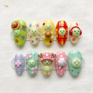 May include: A set of ten press-on nails featuring 3D character embellishments, including a green figure with a straw hat, a figure with red headphones, and a figure wearing a pink hat with antlers, alongside various patterns like plaid, stars, and flowers.