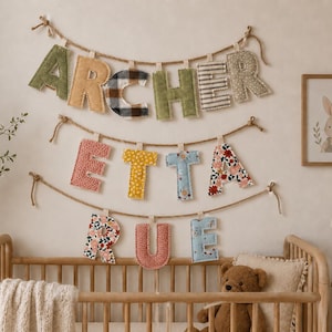 Op de afbeelding: Een muurdecoratie voor de kinderkamer met de naam "ARCHER ETTA RUE" gespeld met stoffen letters. De letters zijn in verschillende patronen en kleuren, opgehangen aan een touw. Een ingelijste konijnenprint en een wieg zijn ook zichtbaar.