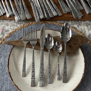 May include: Stainless steel flatware set with decorative handles. The set includes forks, knives, and spoons, arranged on a white plate with a speckled design. Additional pieces are visible in the background on a wooden surface, suggesting a complete dining set.