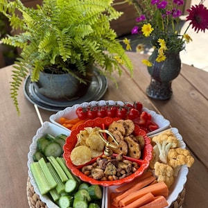 Peut inclure: Un plateau de service compartimenté avec un bol central rouge doté d'une poignée dorée contenant des chips, des bretzels et des noix, entouré de sections blanches remplies de concombres, céleri, carottes, chou-fleur et tomates cerises.