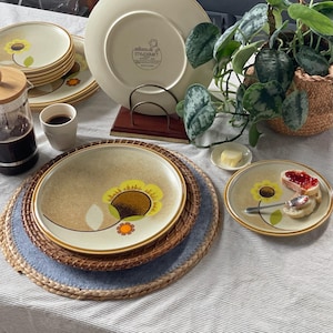 Peut inclure: Ensemble d'assiettes Mikasa Stylecraft avec un motif de tournesol. Les assiettes sont beiges avec un bord marron et un tournesol jaune et marron. Une tasse de café, une cafetière et une petite assiette avec du beurre sont également visibles.