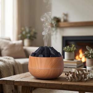 Puede incluir: Un difusor de aceites esenciales con vetas de madera con una pila de objetos negros parecidos al carbón que emiten vapor blanco. El difusor está sobre una mesa de madera rústica, con un ambiente acogedor de salón al fondo.