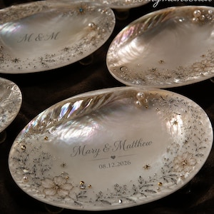 May include: Oval iridescent mother-of-pearl trinket dishes featuring delicate silver floral illustrations, small white pearls, and gold accents. One dish is engraved with the text Mary & Matthew, a heart symbol, and the date 08.12.2026.