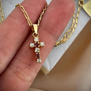 Peut inclure: Un collier à chaîne dorée avec un pendentif en forme de croix orné de pierres blanches transparentes et de pierres rouge foncé alternées. Le pendentif est suspendu à une fine chaîne à maillons, avec une étiquette rectangulaire portant le texte PLAQUE OR.