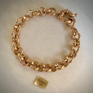 Peut inclure: Un bracelet à maillons dorés composé d'anneaux polis et d'un fermoir à ressort circulaire. Une petite étiquette rectangulaire est placée sous le bracelet, affichant clairement le texte gravé PLAQUÉ OR en lettres capitales.