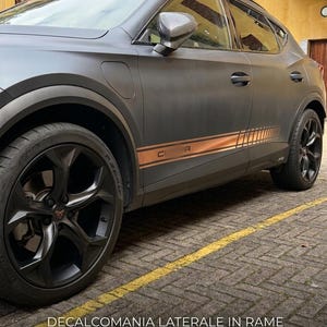 Può includere: Un SUV grigio opaco con cerchi neri e decalcomanie laterali color rame. Il veicolo è parcheggiato su una superficie in mattoni con linee gialle dipinte. La decalcomania laterale ha le lettere "CUPRA".