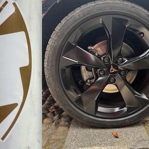 May include: A gold and white decal with a unique shape, next to a black car wheel. The wheel has a black rim with a bronze logo in the center. The tire is black.