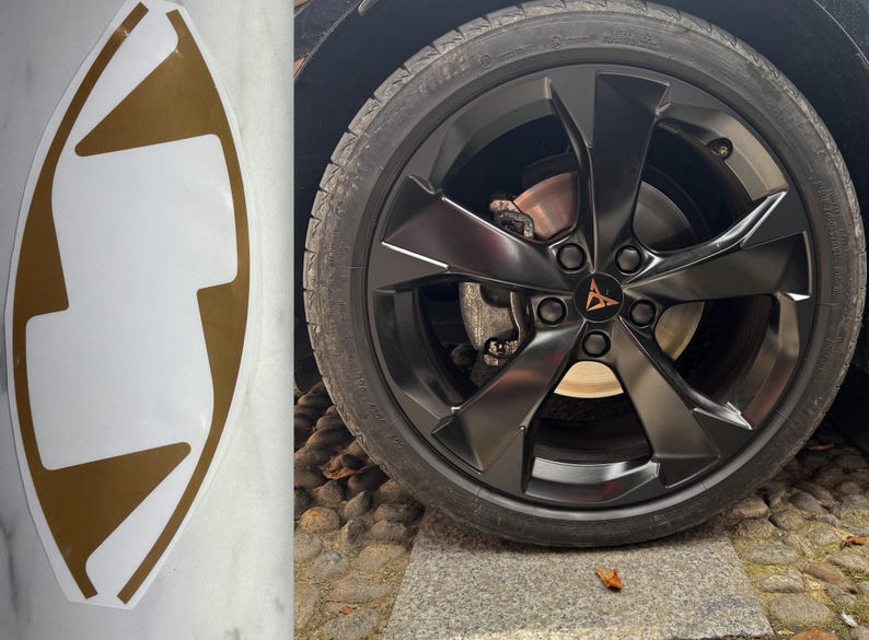May include: A gold and white decal with a unique shape, next to a black car wheel. The wheel has a black rim with a bronze logo in the center. The tire is black.