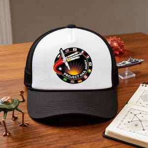 May include: Black and white trucker hat with a white front panel displaying a circular emblem. The emblem features the text "PROJECT HAIL MARY" and a rocket design with flags of various nations. The hat has a black mesh back and a black peak.