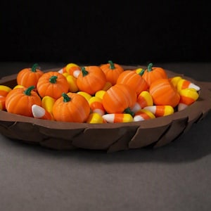 May include: A brown leaf-shaped bowl filled with orange pumpkin-shaped candies and yellow, orange, and white triangular candy corn. The bowl has a textured, layered edge resembling a dried leaf, and the candies are piled inside the center.